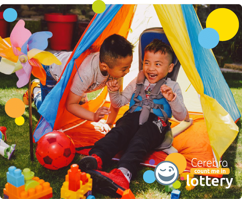 2 young boys, one in a supportive seat, playing in a blue yellow and orange tent, with toys on the grass around them, bottom left, Cerebra count me in lottery logo