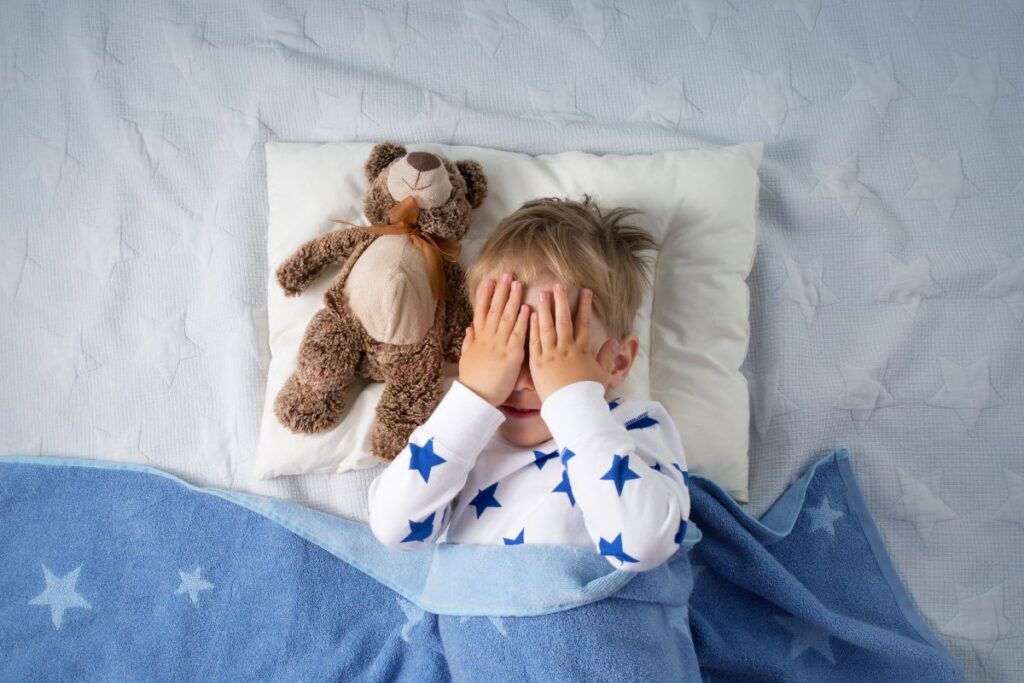 Boy in bed with hands over his eyes next to a teddy