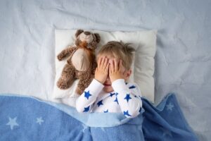 Boy in bed with hands over his eyes next to a teddy