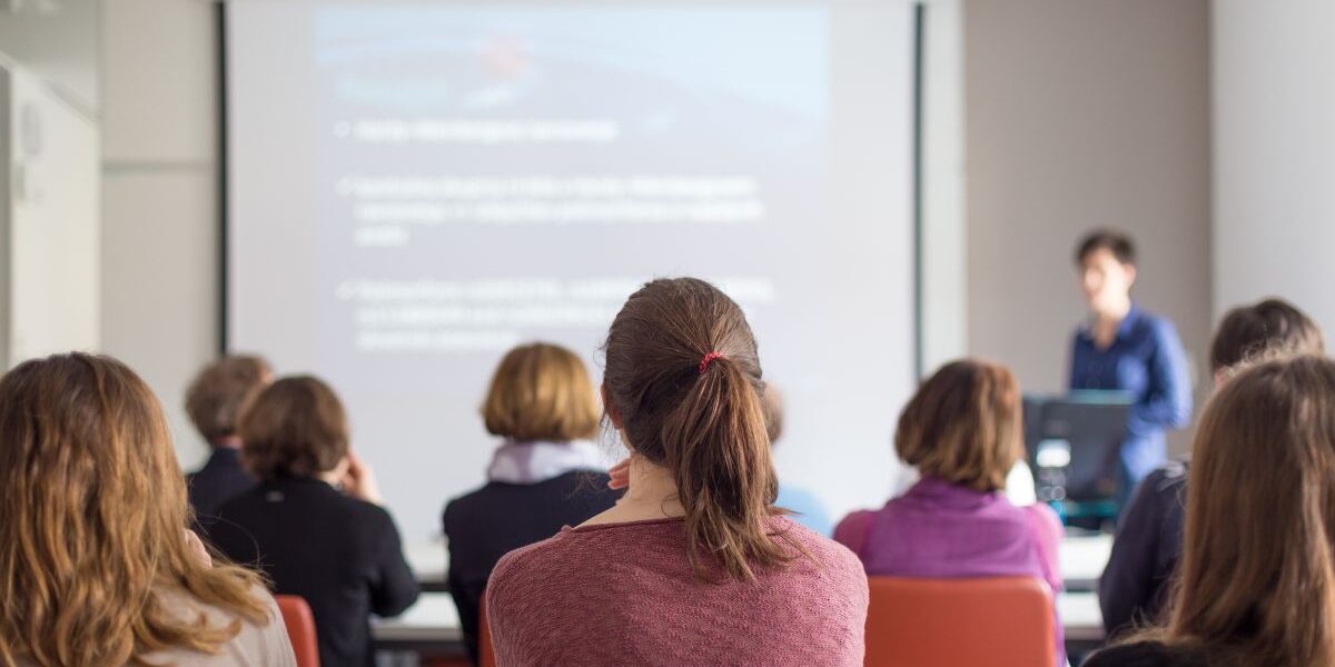 Presentation class 1200px Presenter in the front of a classroom showing a slide show to people with their backs to the camera